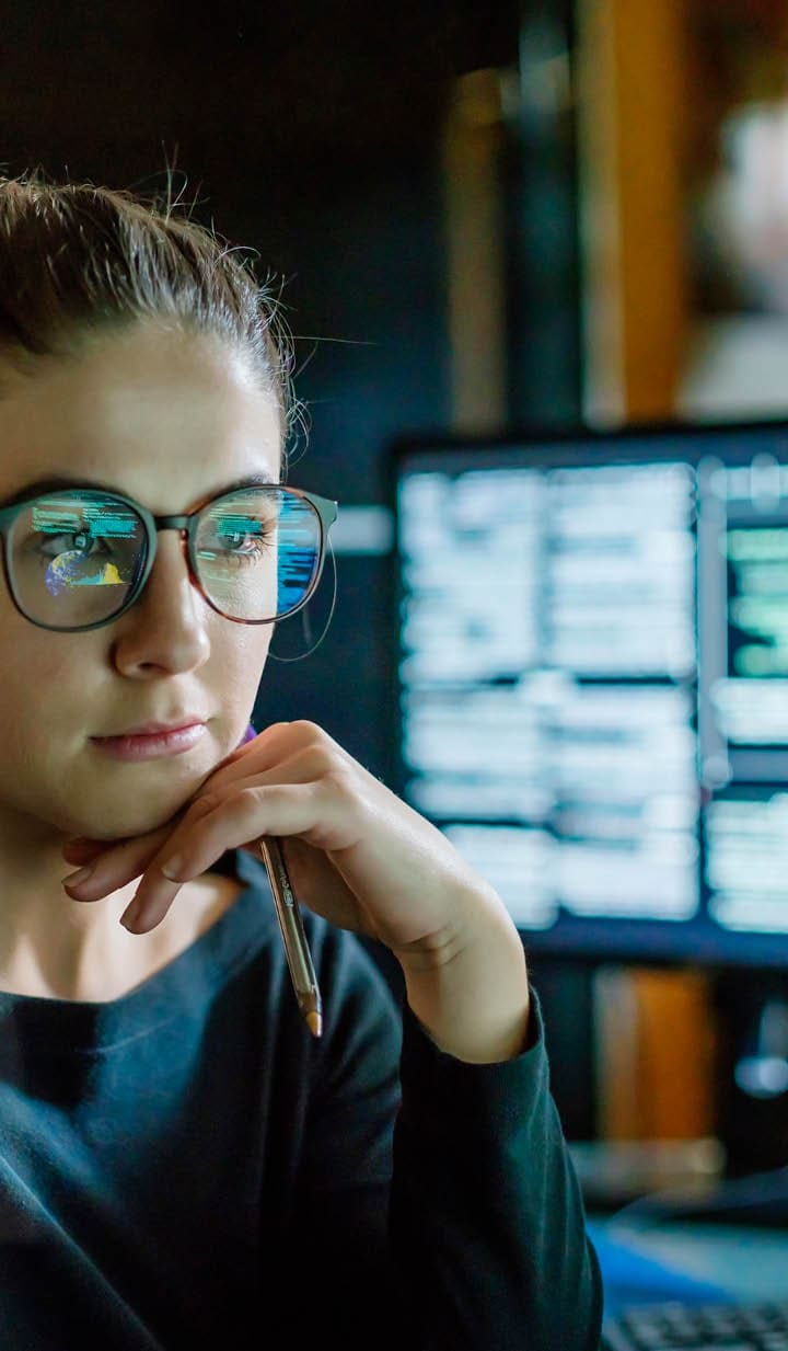 Stock image of a young woman, wearing glasses, surrounded by computer monitors in a dark office. In front of her there is a see-through displaying showing a map of the world with some data.